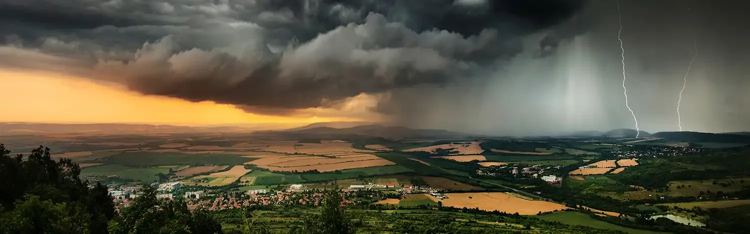 Dramatische Gewitterwolken mit Blitz über ländlicher Landschaft, goldenes Sonnenlicht am Horizont kontrastiert mit dunklem Himmel.
